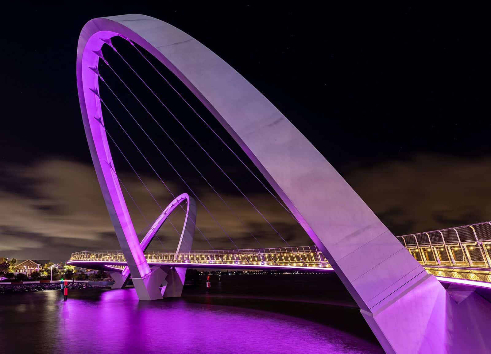 The curved Elizabeth Quay Bridge lit in purple at night, reflected in the water with city towers behind